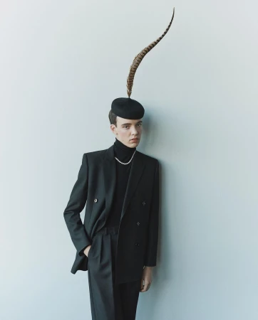 A person in a dark suit with a turtleneck and feathered hat leans against a white wall, photographed in soft, even lighting.