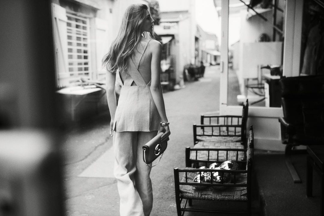 Monochrome shot of a woman in sleeveless top and wide-legged pants walking down an alley, holding a purse, in a fashion editorial style.