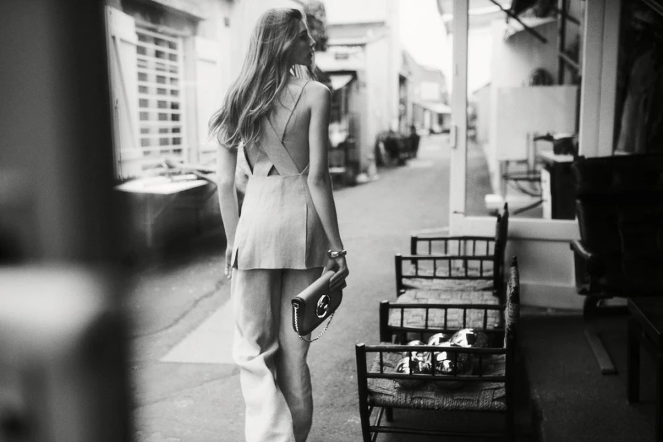 Monochrome shot of a woman in sleeveless top and wide-legged pants walking down an alley, holding a purse, in a fashion editorial style.