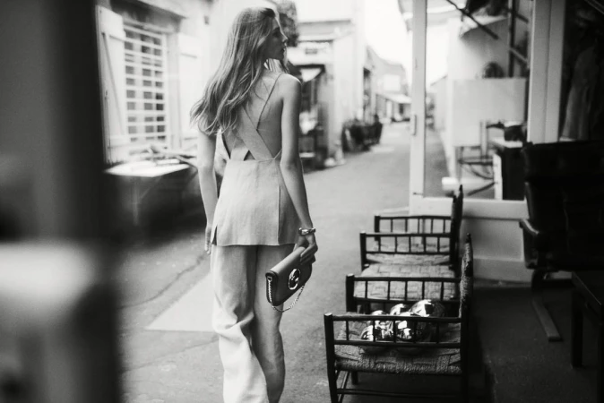 Monochrome shot of a woman in sleeveless top and wide-legged pants walking down an alley, holding a purse, in a fashion editorial style.