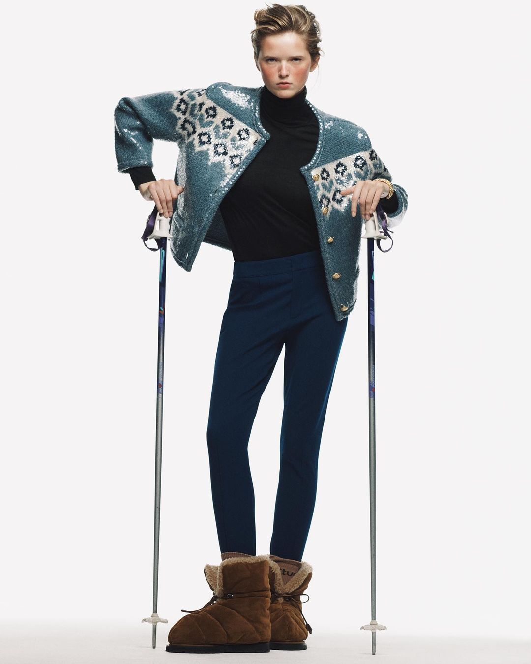 Fashion model poses in a turquoise sequin jacket with nordic pattern, black turtleneck, leggings and winter boots in a studio setting.