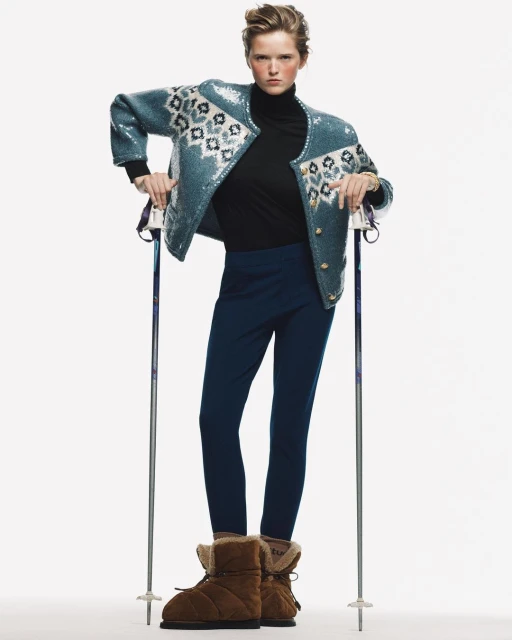 Fashion model poses in a turquoise sequin jacket with nordic pattern, black turtleneck, leggings and winter boots in a studio setting.