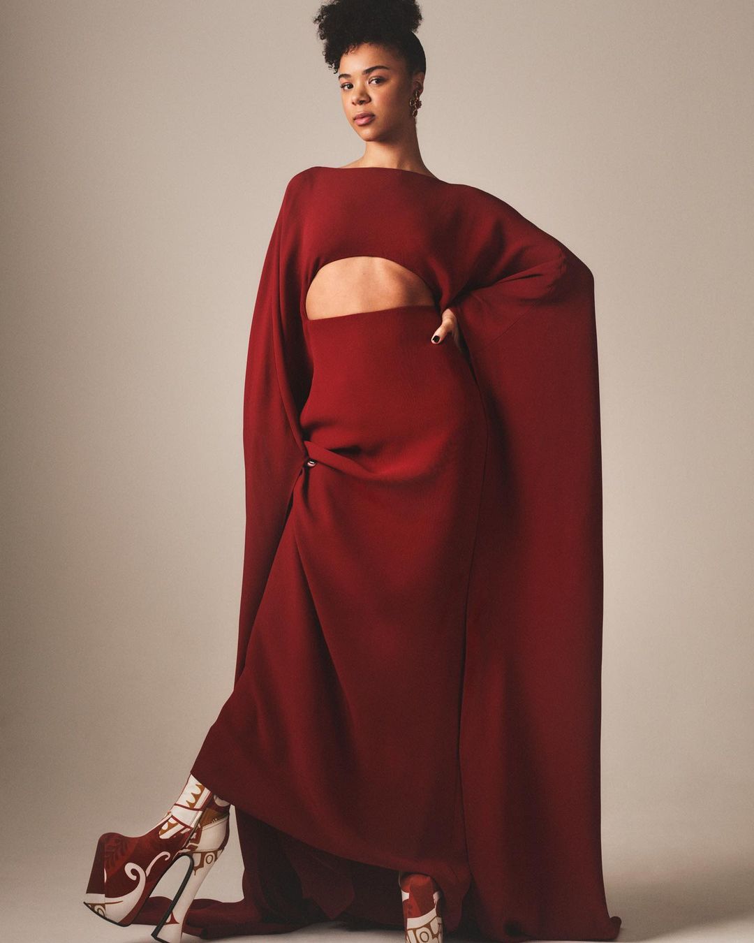 Adult female fashion model poses in a long red dress with a stomach cutout and high platform shoes against a neutral background.