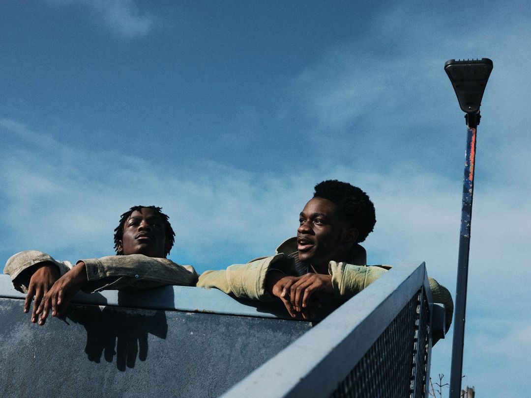 Two African American men in matching jackets lean against a concrete railing under a blue sky with light clouds and a tall metal light pole.