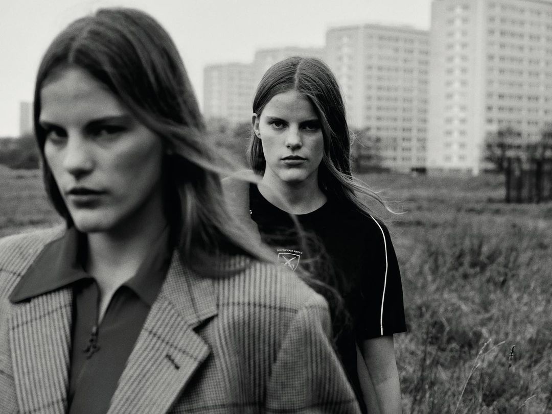 Two young women in black and grey, stand in a field with apartment buildings in the background. The monochrome shot highlights their jackets.