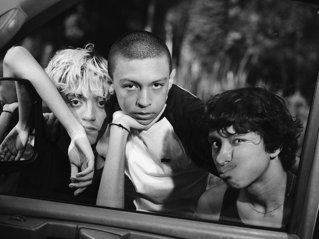 Black and white portrait of three young adults in a car, two looking at the camera and one making a funny face with pursed lips.