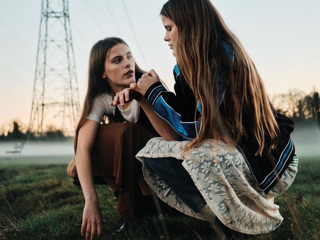 Two young women squatting outdoors in a grassy field near an electrical tower, wearing skirts and casual tops in an outdoor fashion shot.