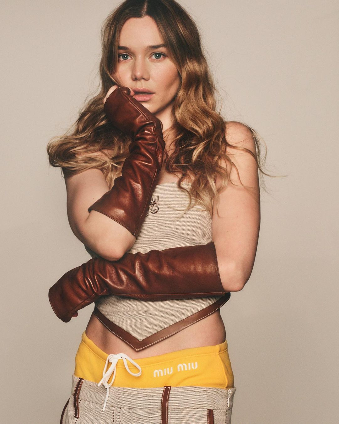 Young woman with long hair poses in studio wearing fingerless leather gloves, tan top, and yellow shorts with logo.