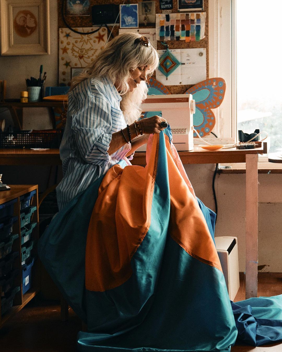 Older woman with gray hair sewing vibrant blue and orange fabric at a sewing machine in a sunlit, home-based craft room.