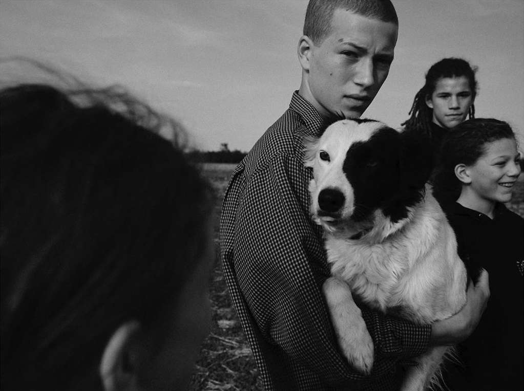Black-and-white portrait of a young man holding a black-and-white dog, with other people standing in the background, taken outdoors.