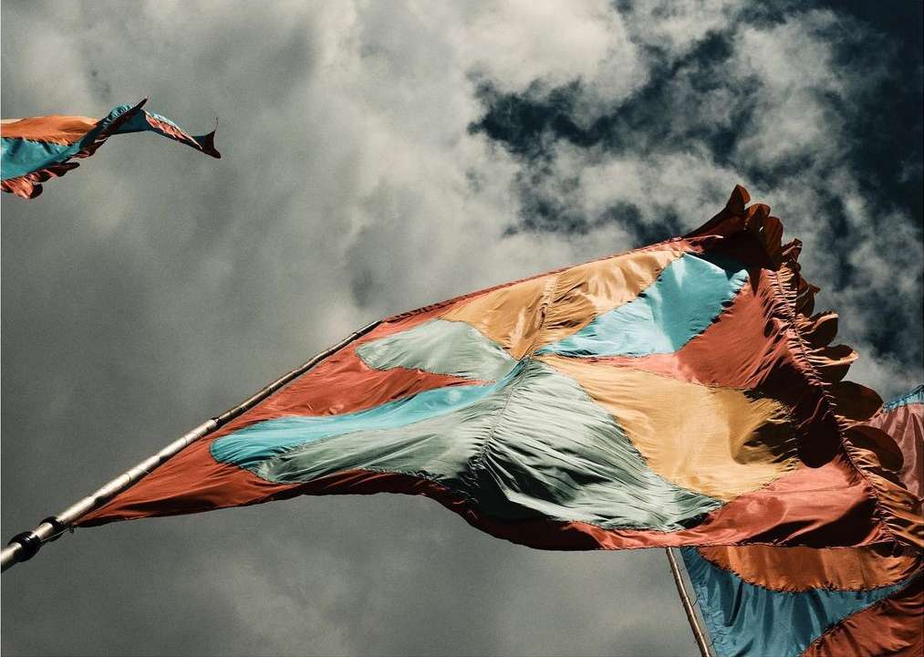 Colorful flags flutter against a moody, cloudy sky, their orange and teal hues providing a striking contrast to the grayscale background.