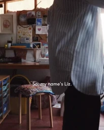 Person wearing a blue and white striped button-down shirt standing in a home workspace with a stool and desk.