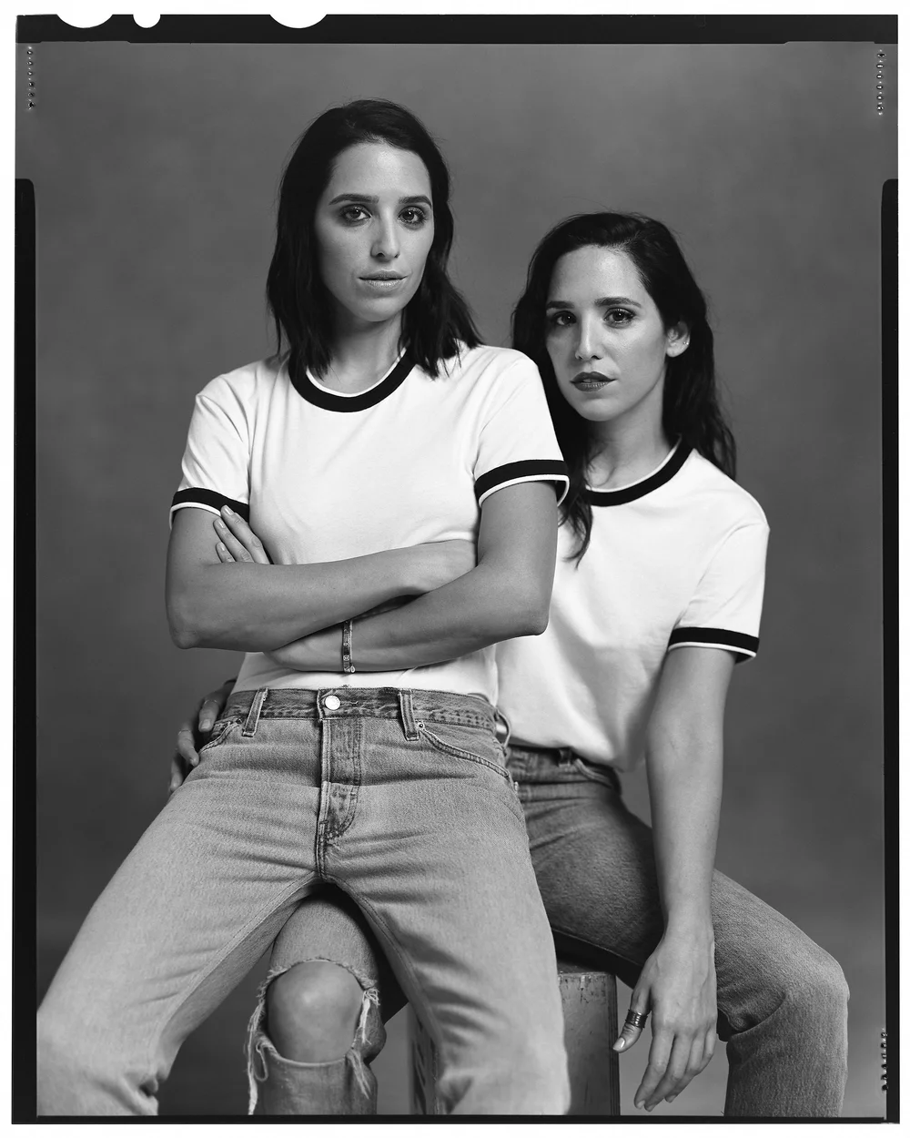 Two women pose in matching white T-shirts and light-wash denim jeans in a black-and-white portrait against a neutral background.
