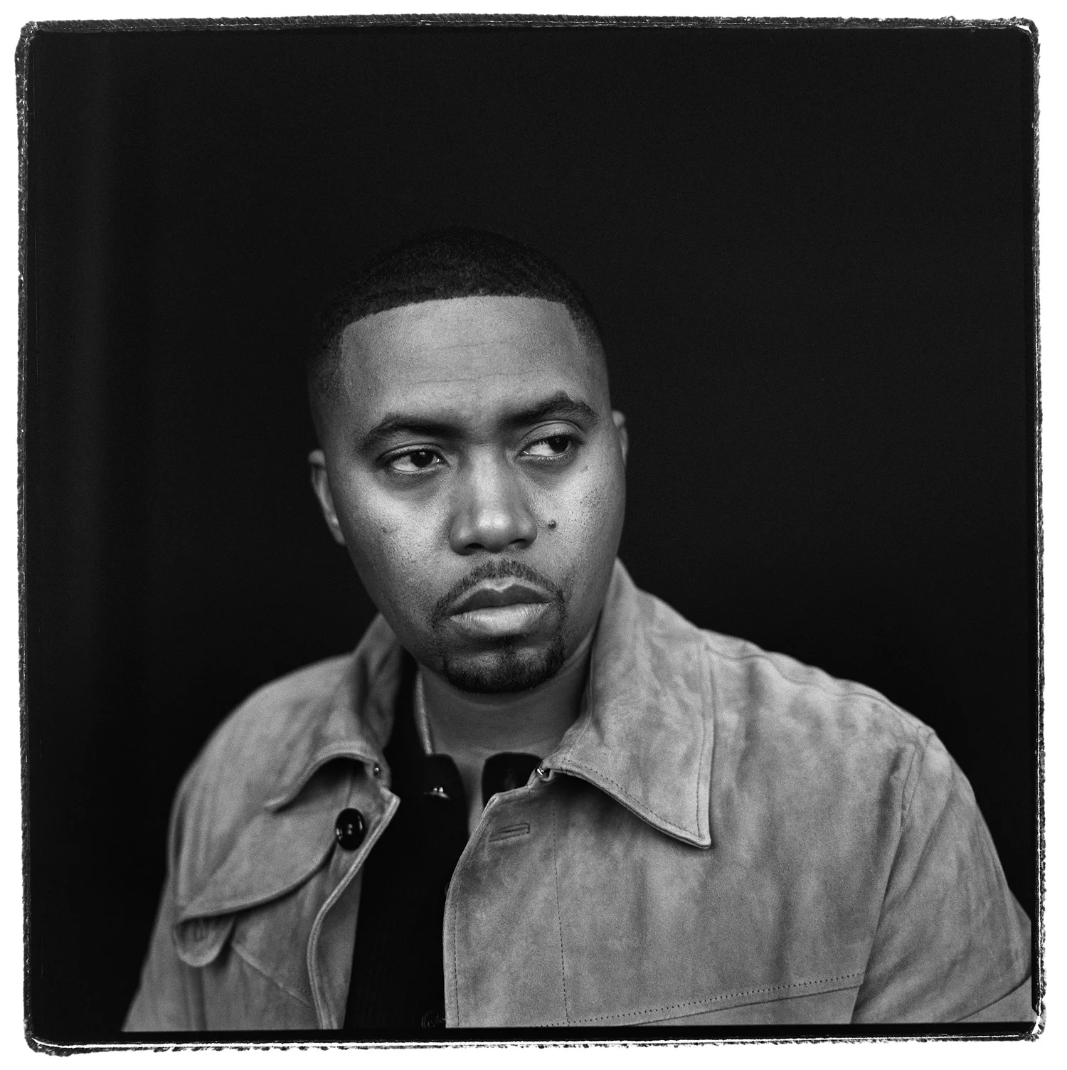 Black and white portrait of Nas wearing a light brown jacket, gazing off to the side against a dark background in an indoor setting.