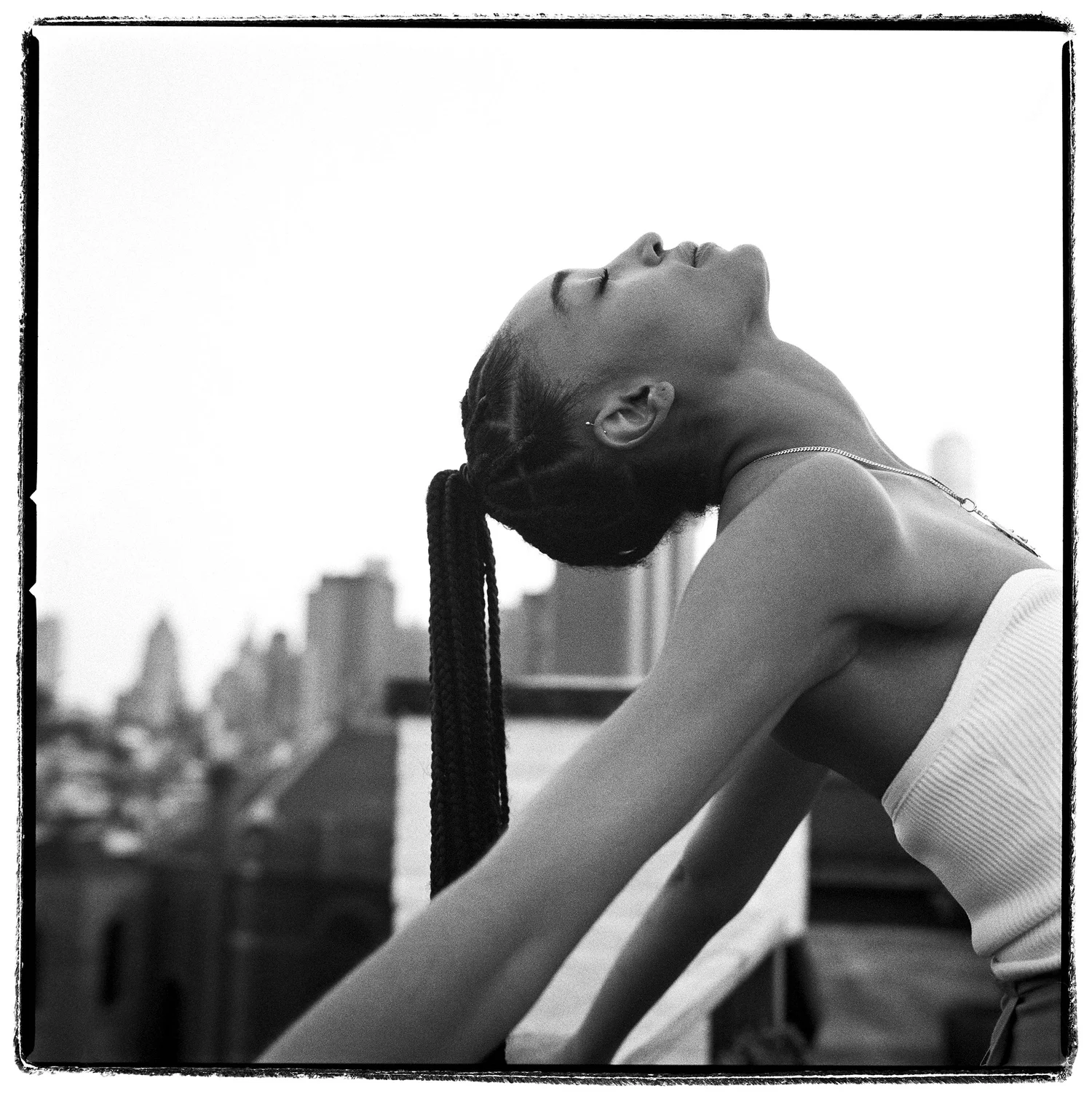 Black and white portrait of a young woman with braided hair, head tilted back, stretching her neck outdoors with a cityscape background.