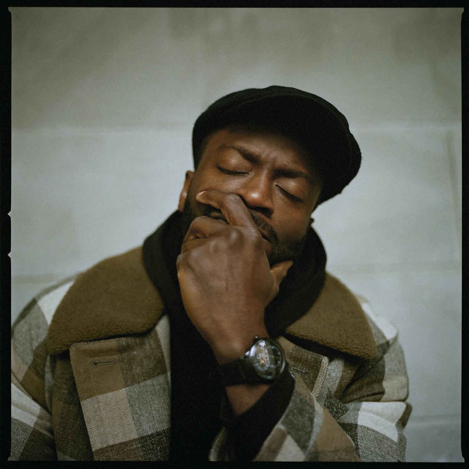 Adult male with dark hair and beard poses indoors wearing a plaid jacket, newsboy cap, and wristwatch against a neutral background.