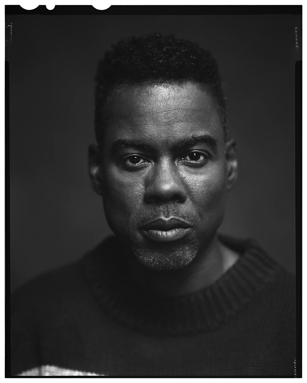 Monochrome portrait of a Black adult male with short hair wearing a dark sweater against a dark background.