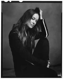 Black and white studio portrait of Olivia Wilde seated with one hand resting on her hair, wearing a black jacket with a white sleeve stripe.