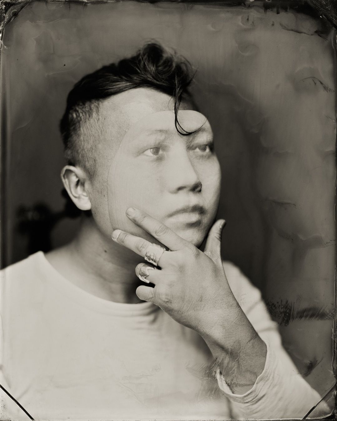 Sepia portrait of an adult with short hair and finger to face, looking upward with a neutral expression in a plain background.