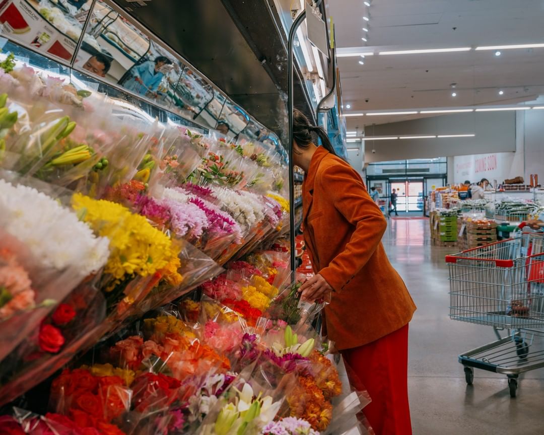 Woman shops for colorful bouquets of flowers inside a bright supermarket with shopping carts visible in the background.