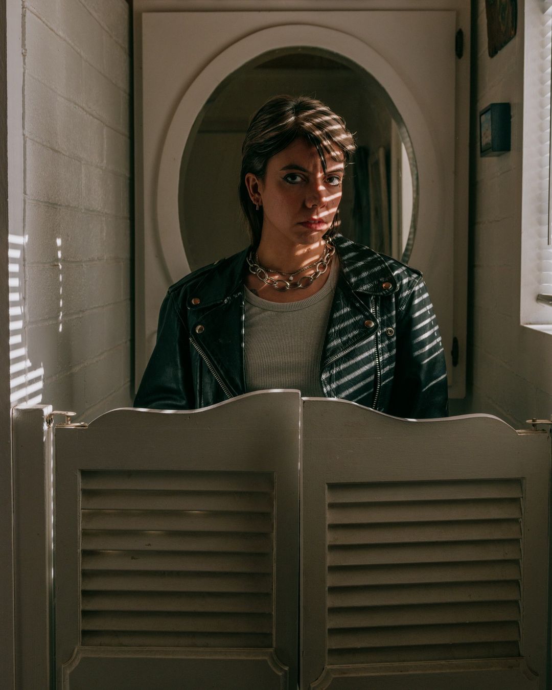 Indoor portrait of a woman in a black leather jacket with a serious expression, featuring striped lighting effects and bathroom saloon doors.