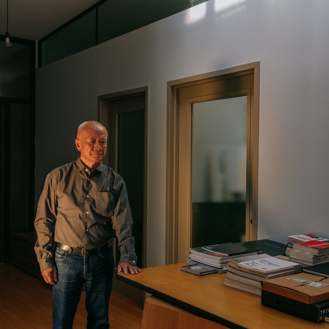 Indoor portrait of an Asian man standing near a desk with books in a room with bright daylight and two doors in the background.