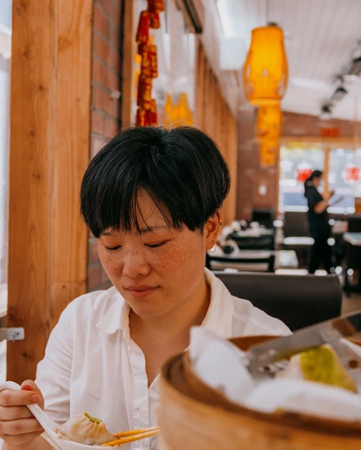 Asian woman in white shirt enjoys dim sum meal with chopsticks and spoon in a bright restaurant with hanging red lanterns.