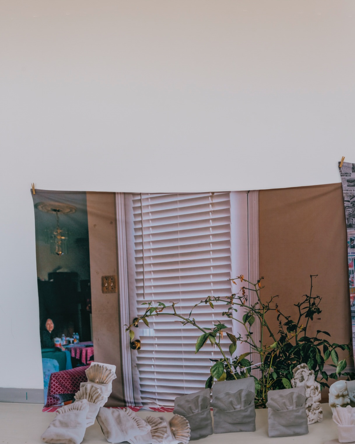 Abstract art installation featuring a tapestry with window and figure, potted plants, and white textured objects against a plain wall.