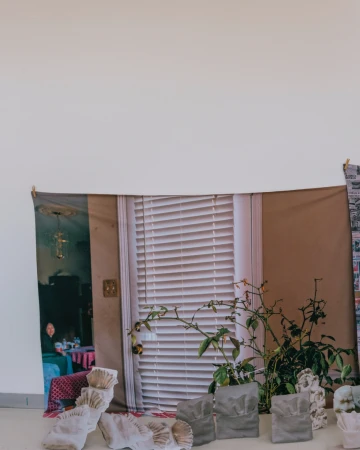 Abstract art installation featuring a tapestry with window and figure, potted plants, and white textured objects against a plain wall.