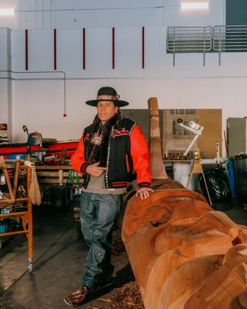 Native American man poses next to a large carved wooden statue inside a woodworking shop, wearing a hat and a custom jacket.