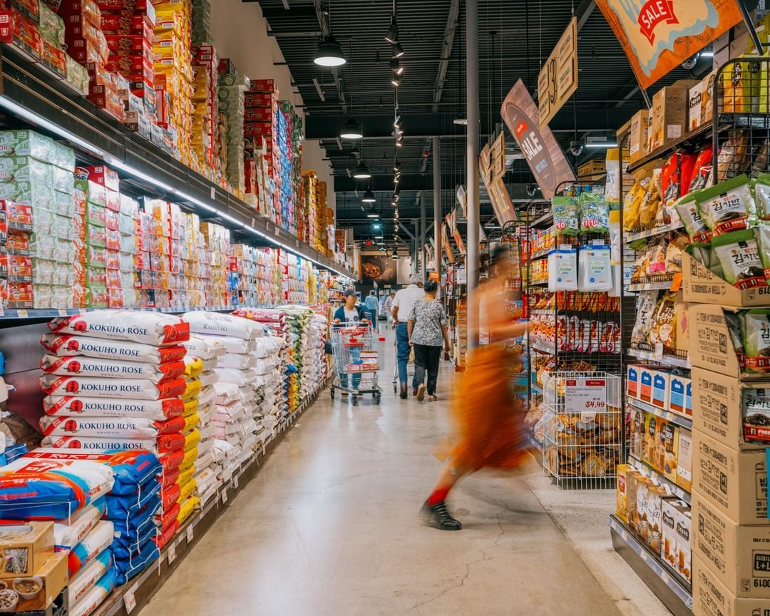 Grocery store aisle with stacks of goods, blurred shoppers and shopping carts. Overhead lights and sale signs visible.
