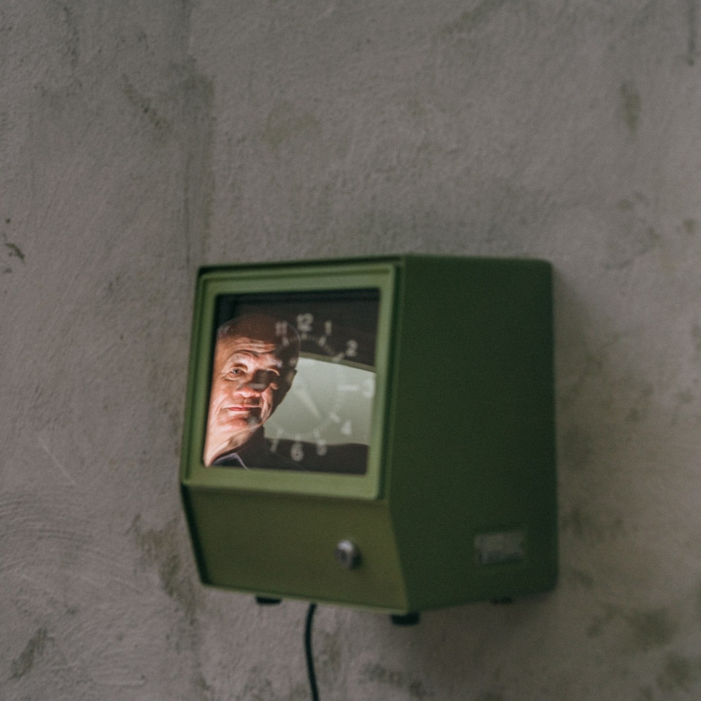 An elderly man's face is reflected in the glass of a vintage, green wall-mounted clock, against a textured grey wall.