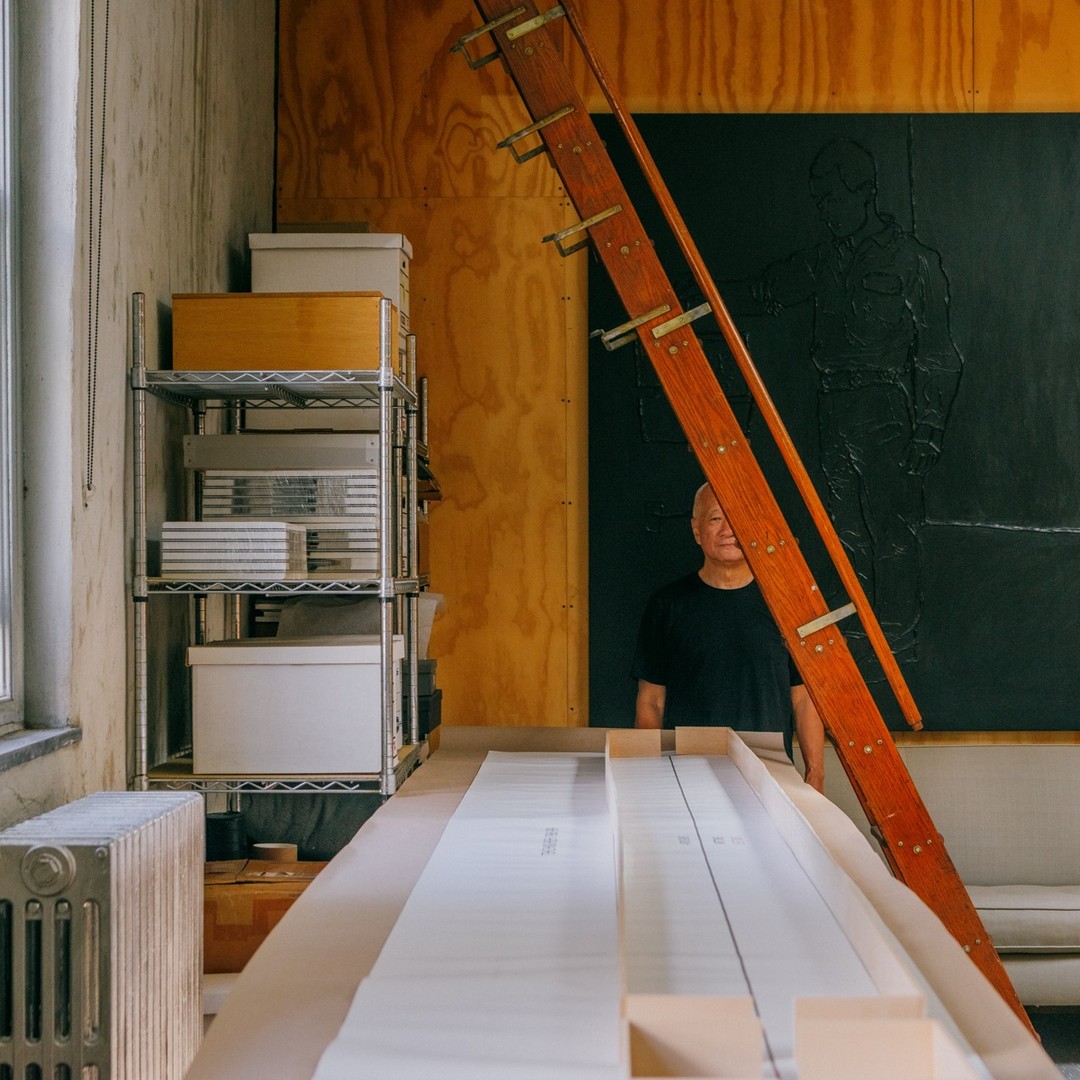 Mature man in black shirt stands in artist's studio with ladder and artwork in the background surrounded by boxes and paper rolls.
