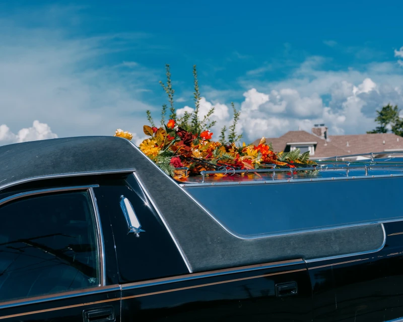 Close-up of a black hearse adorned with colorful flowers on its roof, set against a bright blue sky with scattered clouds and a residential building.