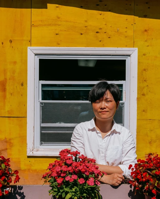 Asian woman in white shirt poses at white-frame window of yellow house exterior with red geraniums in sunlight, creating a warm, inviting scene.