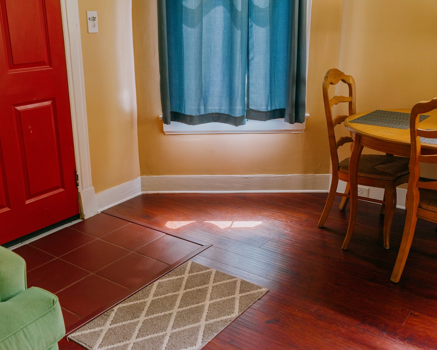 Interior shot of a dining room with a wooden table and chairs, a red door, blue curtains, and a neutral-toned rug on a hardwood floor.