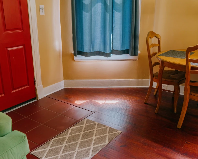 Interior shot of a dining room with a wooden table and chairs, a red door, blue curtains, and a neutral-toned rug on a hardwood floor.