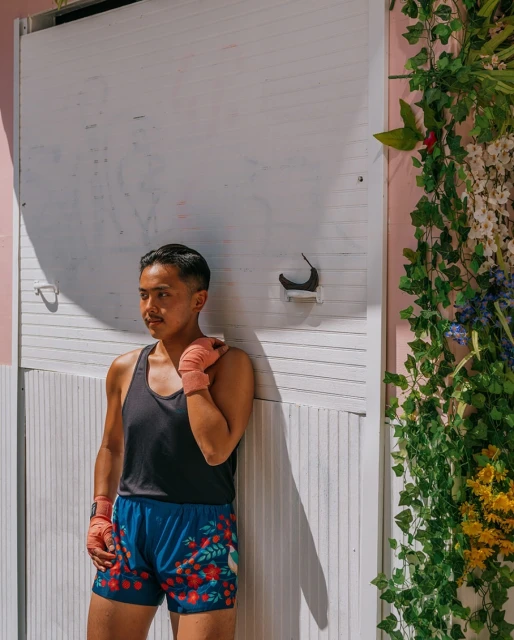 Athletic man with hand wraps leans against a white wall with floral decorations, wearing a tank top and floral shorts in outdoor light.