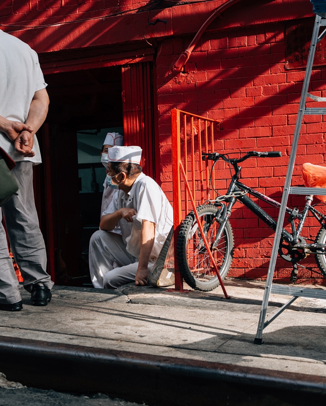 Two adults sit in front of red brick building; one man stands nearby. A black bicycle and a metal ladder are near the wall.