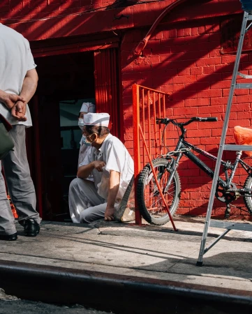 Two adults sit in front of red brick building; one man stands nearby. A black bicycle and a metal ladder are near the wall.