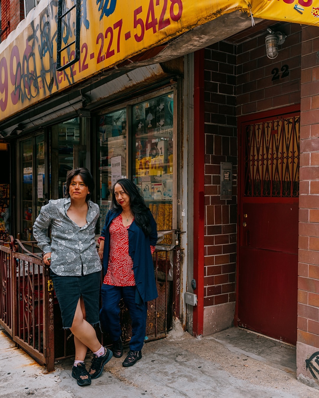 Two adult women stand together on a city sidewalk in front of a store with yellow signage, with a brick wall and door to the right.