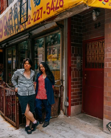 Two adult women stand together on a city sidewalk in front of a store with yellow signage, with a brick wall and door to the right.