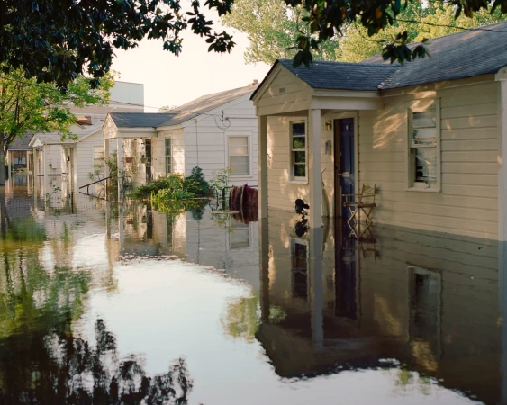 Flooded residential neighborhood showing houses partially submerged in water, reflecting sky and trees. A natural disaster image.