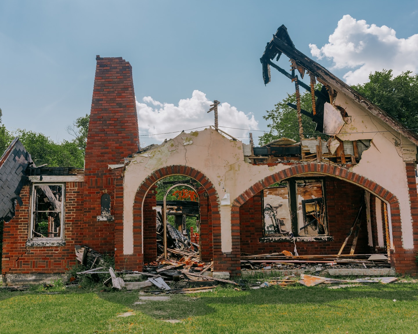 Exterior of a dilapidated house with fire damage shows brick chimney, walls, arches, and debris against a blue sky with scattered clouds.