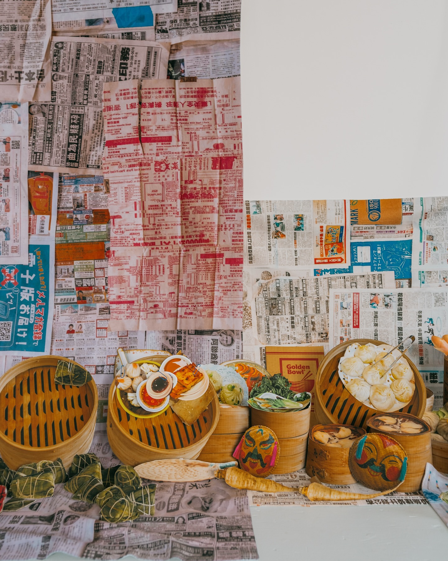 Overhead shot of bamboo steamer baskets filled with dim sum against a background of newspaper clippings and a blank white canvas.