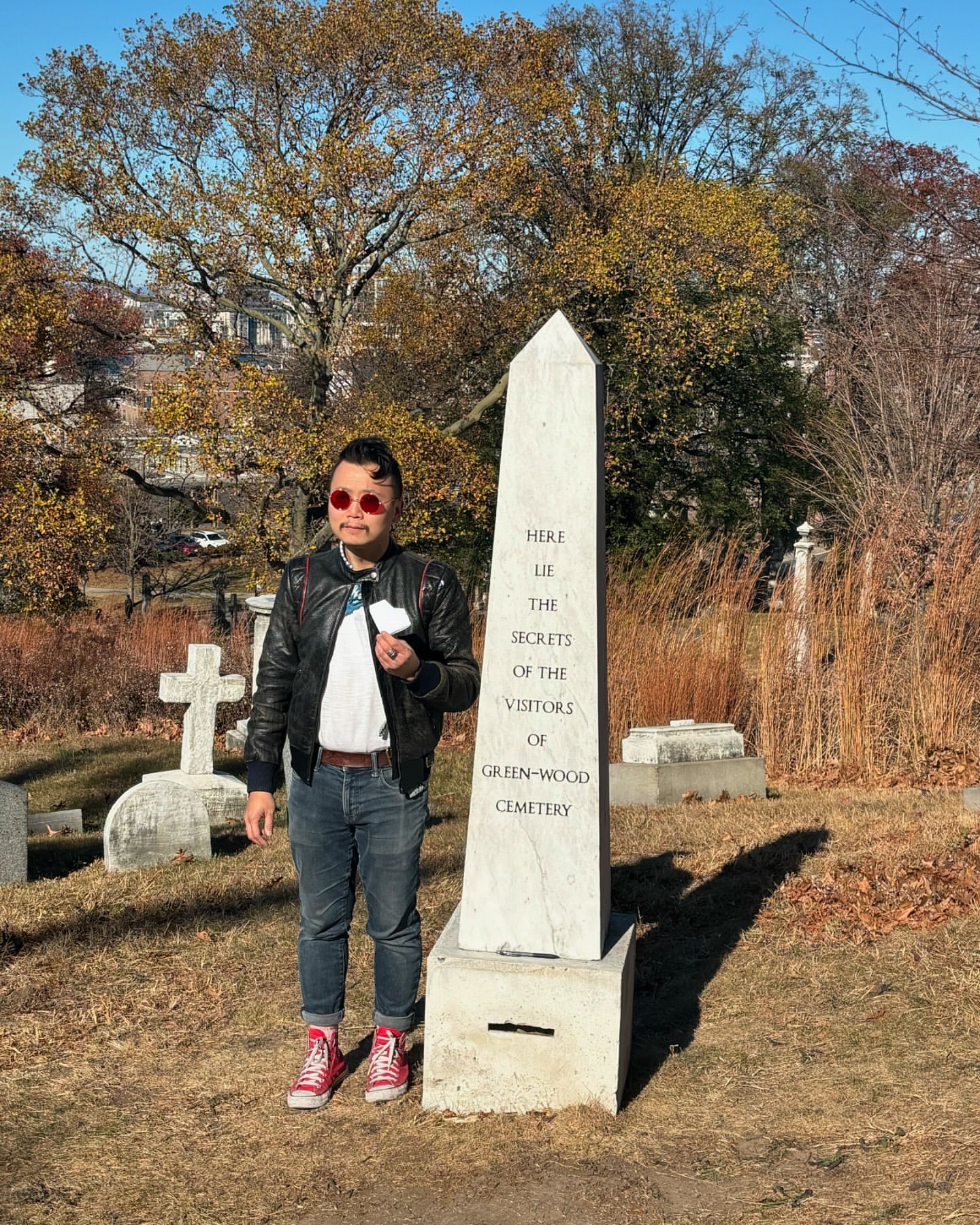 Adult male poses near a marble obelisk in an outdoor cemetery setting with trees and other headstones visible in the background.