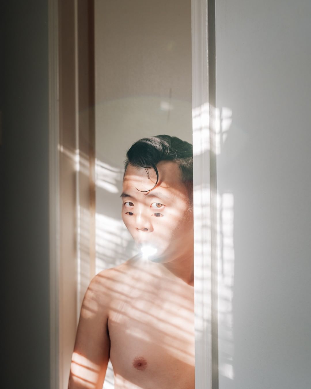Portrait of an Asian man standing indoors with sunlight and shadows across his body and face, highlighting his dark, wet hair.