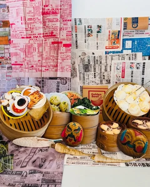 Artful food still life of varied Asian dumplings and dim sum in bamboo steamers against a backdrop of vintage newspaper clippings.