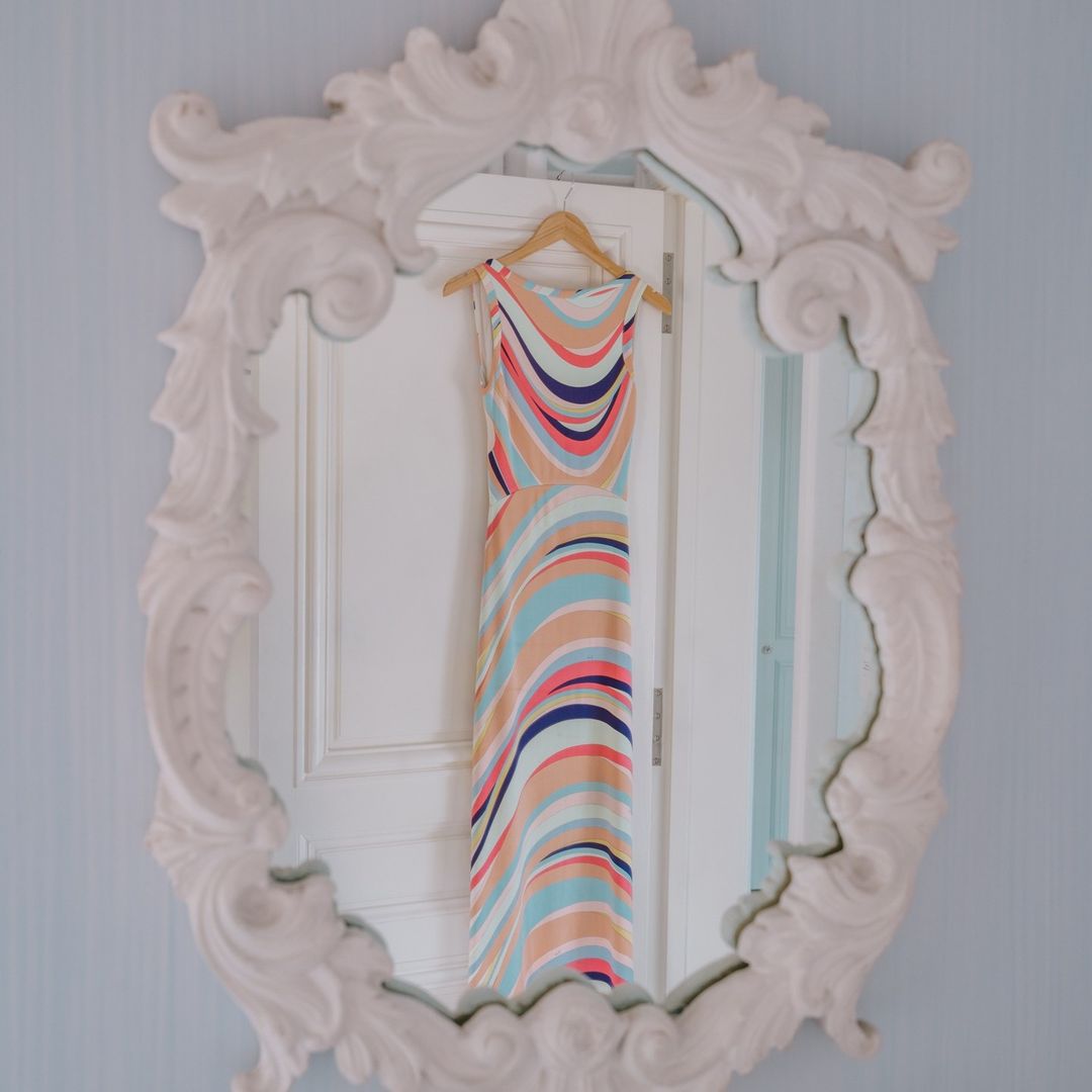 Elegant sleeveless dress with colorful stripes hanging on a wooden hanger, reflected in a decorative white mirror on a blue wall.