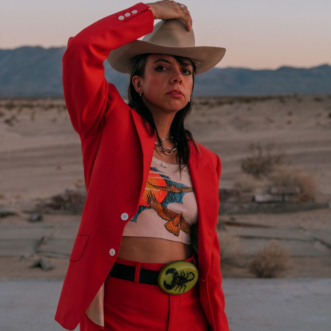 Stylish woman in red suit, cowboy hat poses in desert landscape with mountains at sunset. Fashion, style, desert aesthetic.